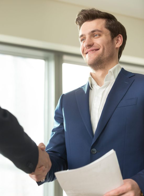 Confident smiling businessman with documents in hand handshaking with partner at business meeting in office. Presentable financial consultant welcoming client, congratulating investor with good deal